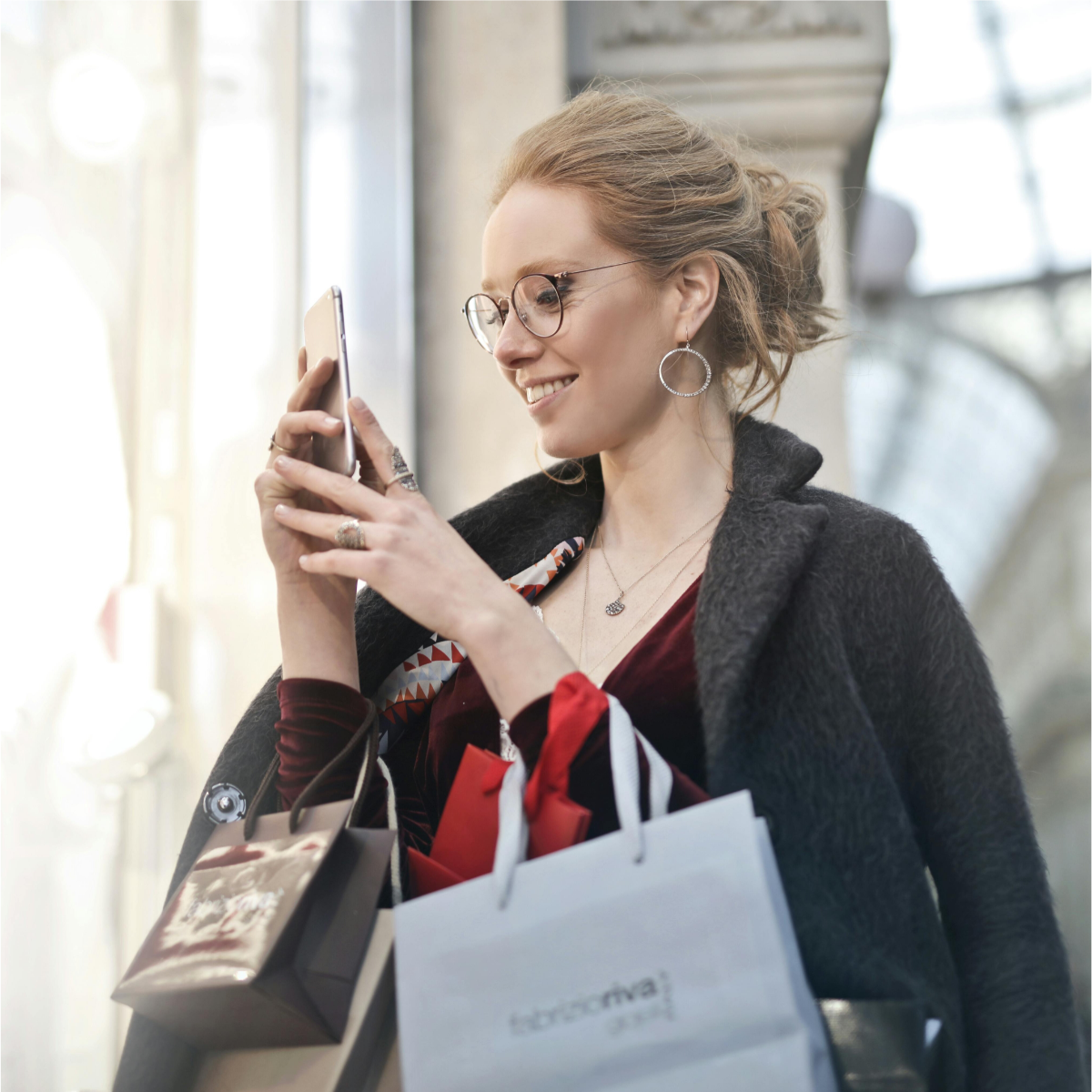 Woman wearing glasses with lots of shopping bags draped on her arms looks at her mobile device and smiles.
