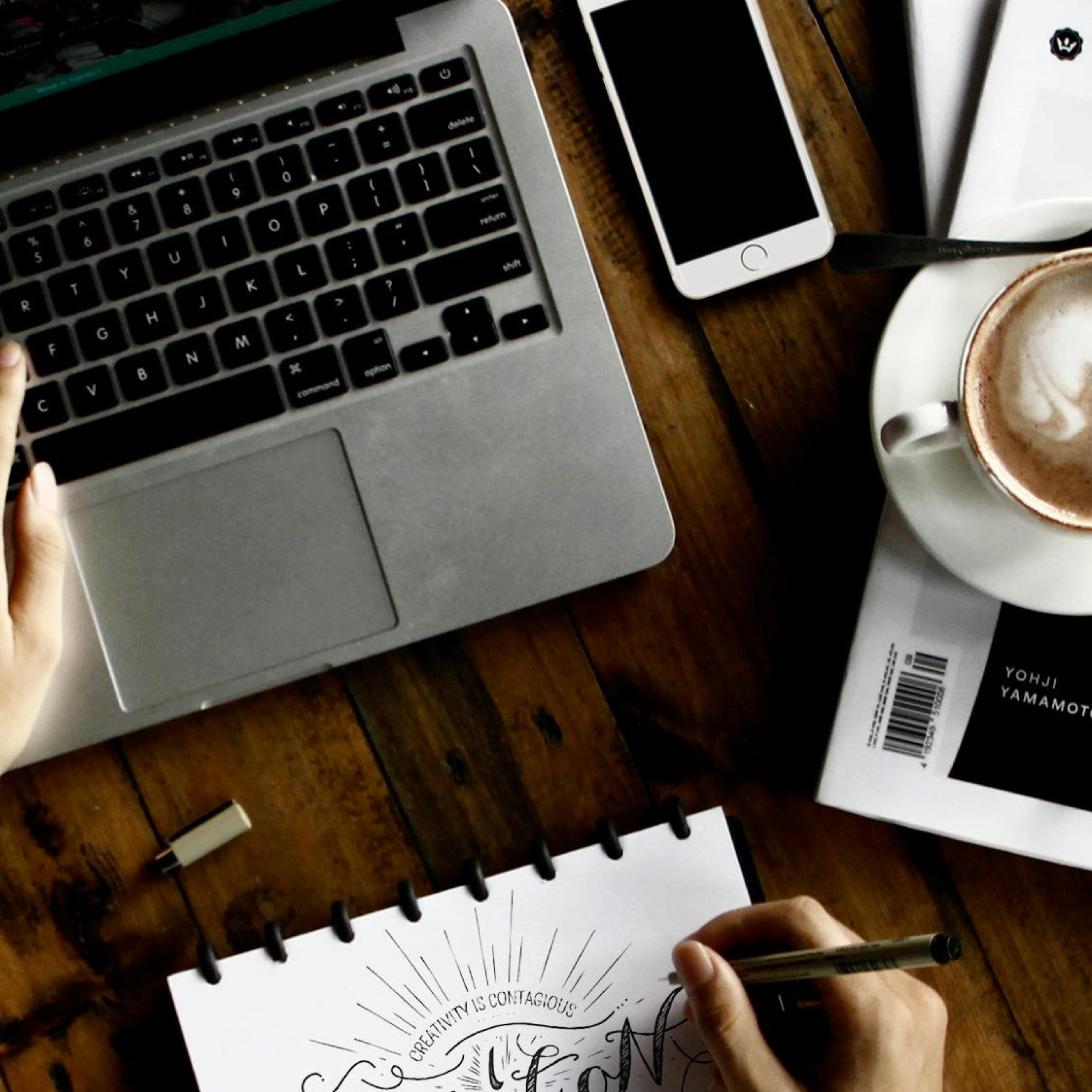 Desk shown with open laptop, cell phone, coffee mug, and a person writing on a notepad. The notepad says Creativity is contagious.