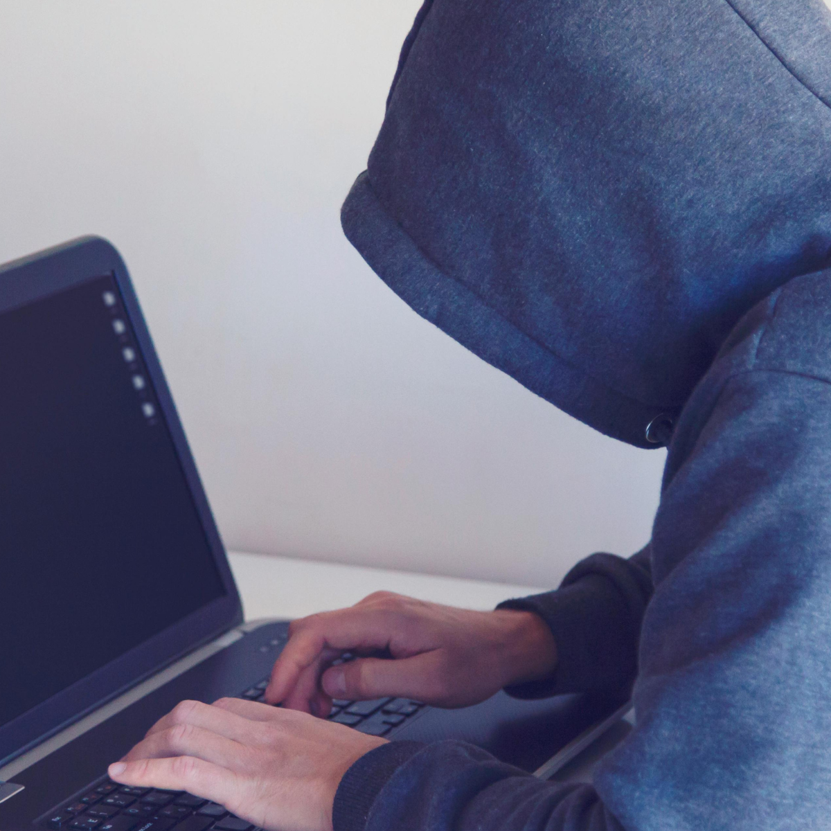 A hacker with a hoodie covering his face is at a small white table working on a laptop.