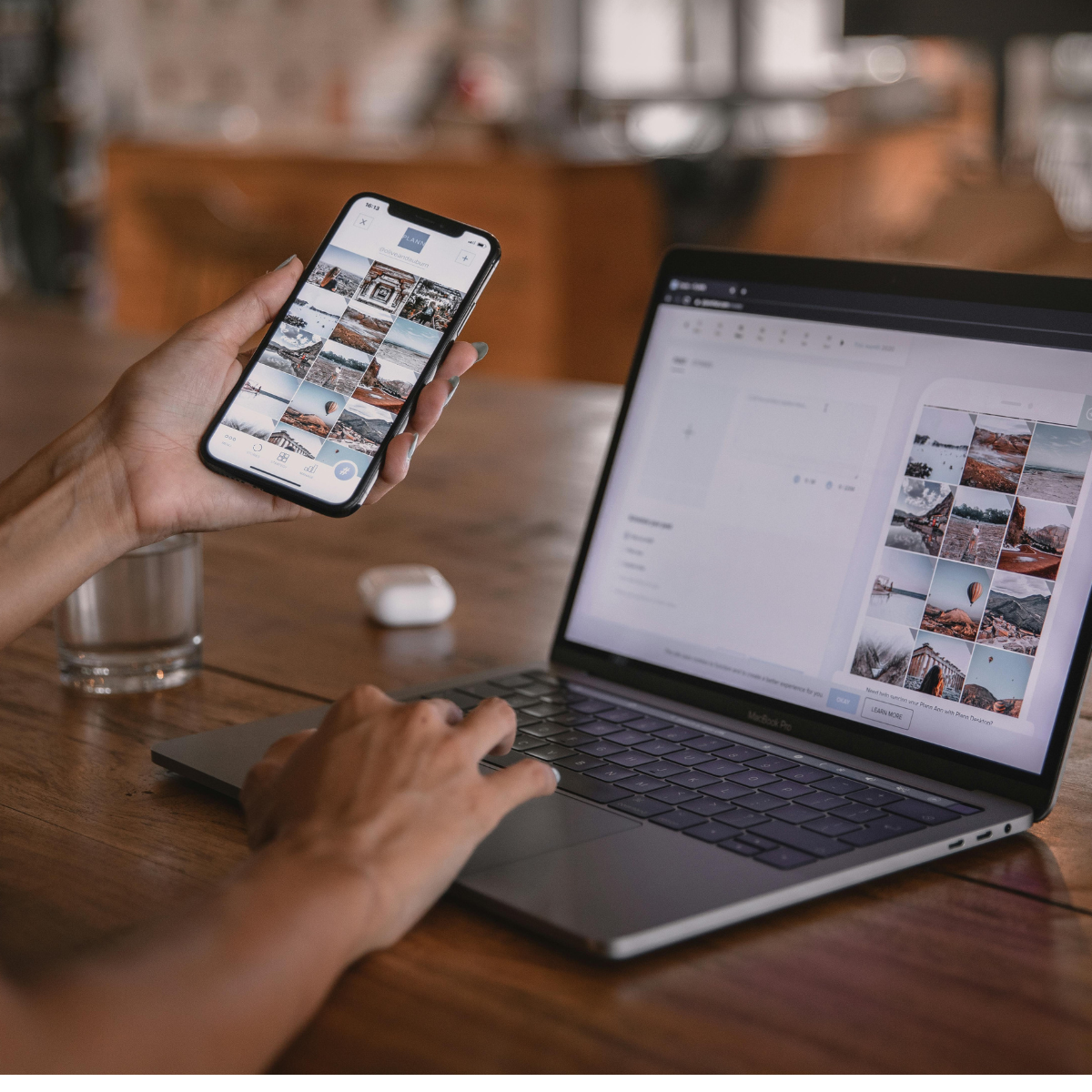 Image of woman's left hand holding smart phone while right hand types on a laptop. A glass and earpod case rest to the left on the wooden table.