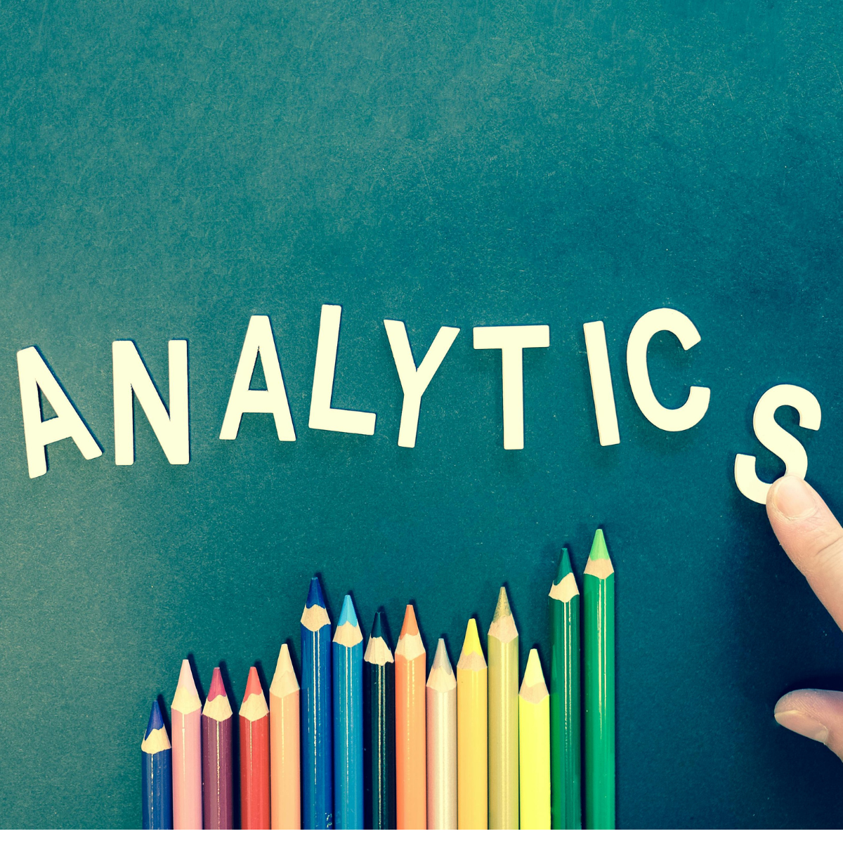Green background with the letters for ANALYTICS in white. An index finger is adjusting the S. A rainbow assortment of colored pencils are lined up along the bottom of the image.
