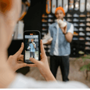A white woman is recording a black man using a cell phone as he stands in front of a wall of shoes and holds one pair while talking.