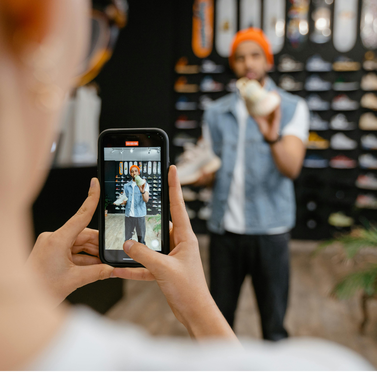 A white woman is recording a black man using a cell phone as he stands in front of a wall of shoes and holds one pair while talking.