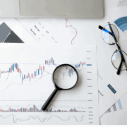 Graphs and other analytics printouts in red, blue, and black on white paper rest on a white table with a pair of black eyeglasses and a black magnifying glass.
