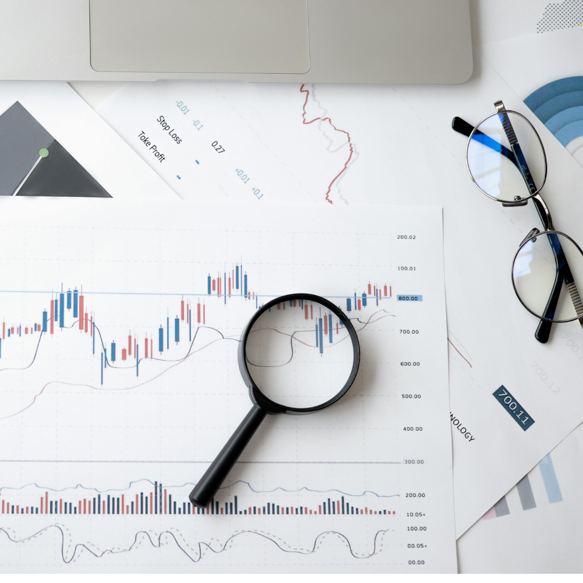 Graphs and other analytics printouts in red, blue, and black on white paper rest on a white table with a pair of black eyeglasses and a black magnifying glass.