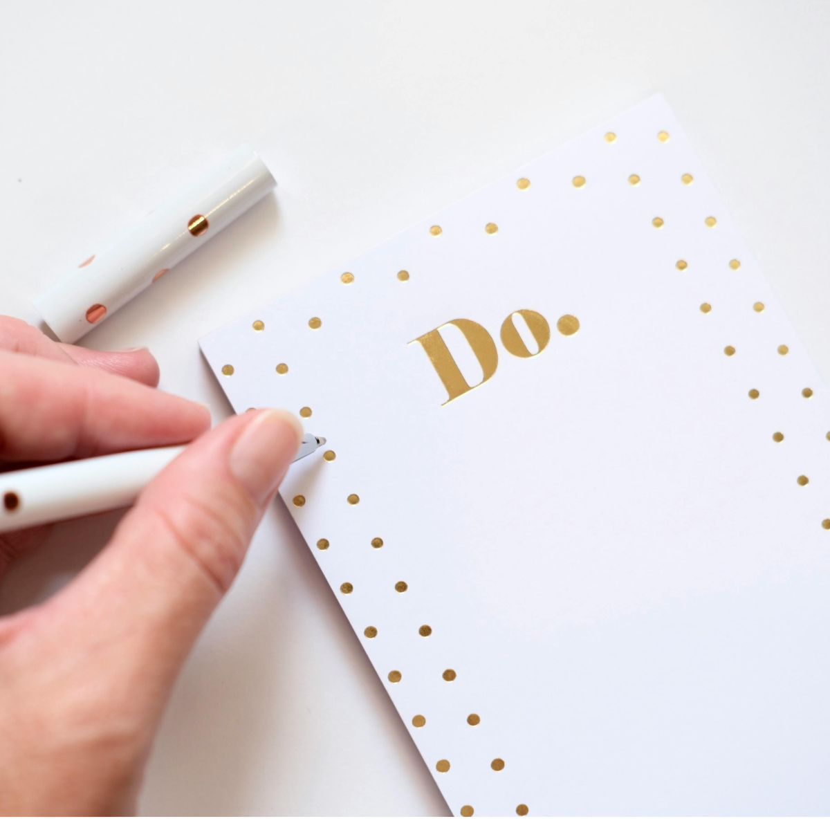 White notepad with gold word DO. and polka dots rests on a white surface, and a white person's left hand holds a white pen with gold dots.