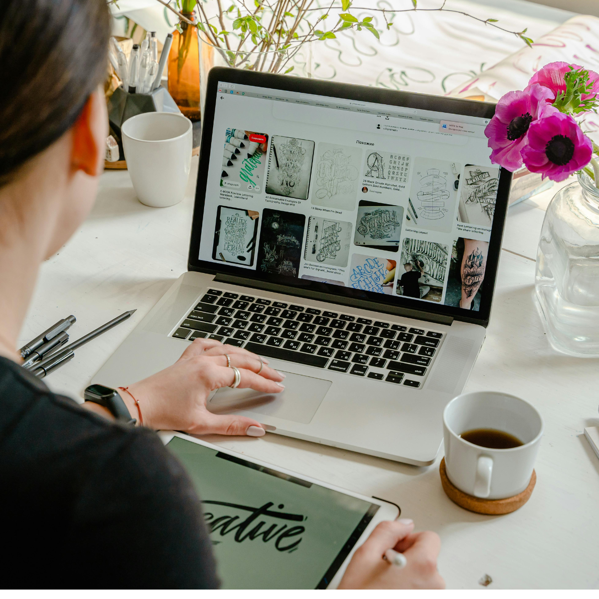 Woman sits at desk working on laptop with a notebook, pens, coffee, and a vase of pink flowers nearby. She appears to be browsing design elements or photos.