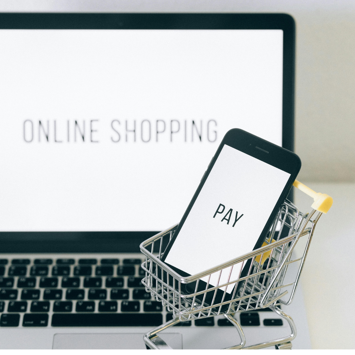 A laptop screen reads "online shopping" and a tiny toy shopping cart sits in the foreground holding a mobile phone with the word "pay" on the screen.