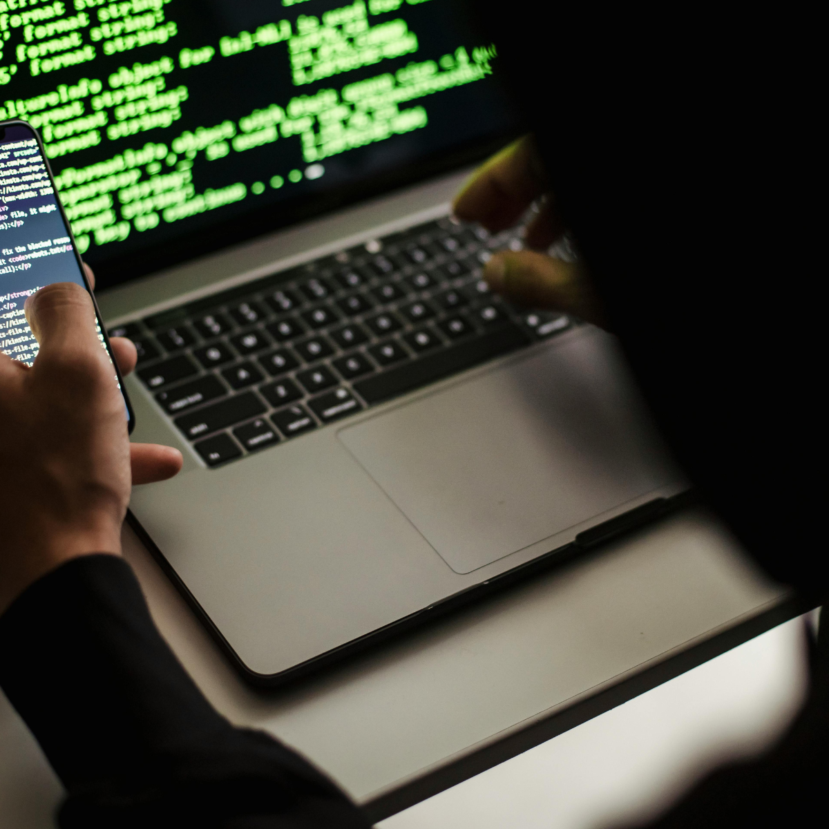 A silver and black laptop rests on a white surface while a person in a black hoodie leans over it typing and also holding a mobile phone. Computer code appears on each screen.