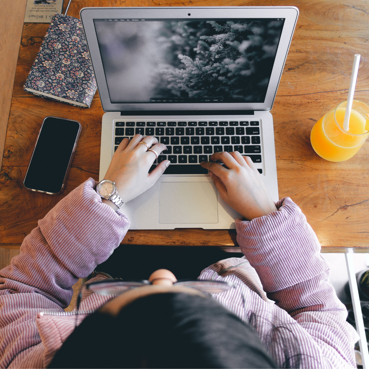 A woman sits and types on a laptop resting on a wooden table, with a notebook, cell phone, and glass of orange juice with a straw nearby.