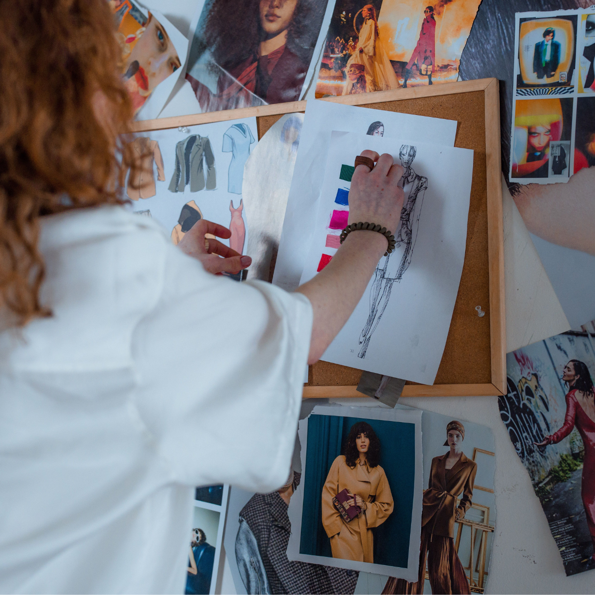 A woman stands at a bulletin board and arranges images and drawings for a fashion business.